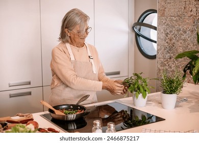 Smiling senior gray haired woman at home cooking vegetables in the pan adds some herbs. People in the kitchen preparing dinner. An elderly woman works in the kitchen - Powered by Shutterstock