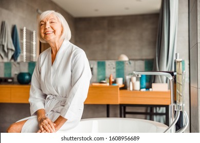 Smiling senior lady in bathrobe sitting on the edge of bathtub stock photo