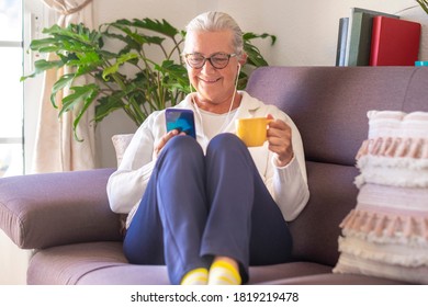 Smiling Senior Lady Sitting On Sofa At Home Using Her Smart Phone For Chatting With Friends Or Family - Woman White Hair, Glasses Wearing Earphones - Active Social And Tech People