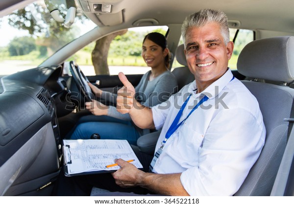 smiling senior male driving instructor in a car\
with learner driver giving thumb\
up