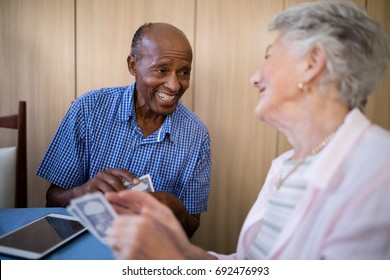 Smiling Senior Male And Female Friends Playing Cards At Table In Nursing Home