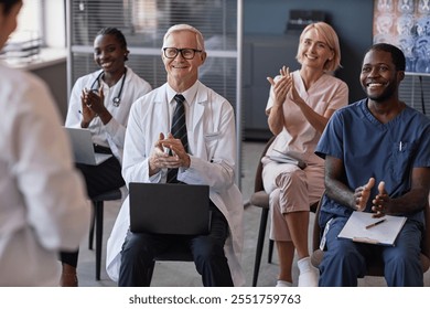 Smiling senior male head physician supporting speaker with applause sitting in conference room with diverse team of doctors during colleagues presentation - Powered by Shutterstock