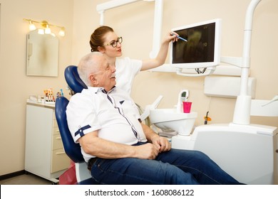 Smiling Senior Man 70-75 Years Old On Review Of Young Woman Dentist, Durring A Medical Consultation At Dental Office, Explaining X-Ray Results To Man. Dentistry, Medicine And Healthcare Concept.