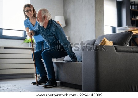 Smiling senior man with female healthcare worker. Home carer supporting old man to stand up from the chair at care home.. Female caregiver helping senior man get up from couch in living room - Indoors 商業照片 © 