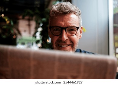 Smiling senior man in glasses reads the newspaper at a cozy cafe, enjoying the latest news and stories, reflecting on community and urban life - Powered by Shutterstock