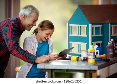 Smiling Senior Man And His Granddaughter Having Fun Looking At A IPad Together In The Garage.