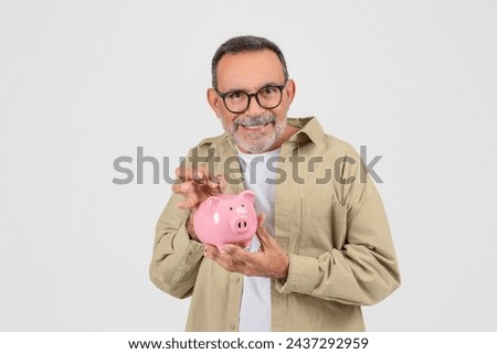 Smiling senior man holding and inserting coin into pink piggy bank, happy elderly gentleman representing smart savings and financial planning, standing on white background in studio, copy space