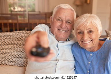 Smiling senior man pointing a remote while sitting with his wife on a couch in their living room watching television together