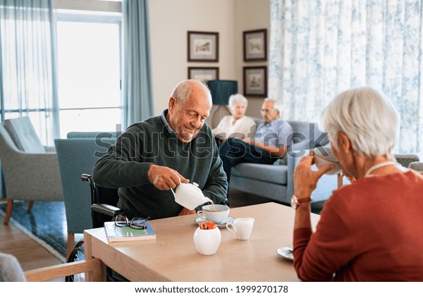 Smiling senior man pouring tea in cup from teapot\
in care facility with wife sitting at table in the common area.\
Elderly man in care centre sitting on wheelchair serving tea during\
a visit.