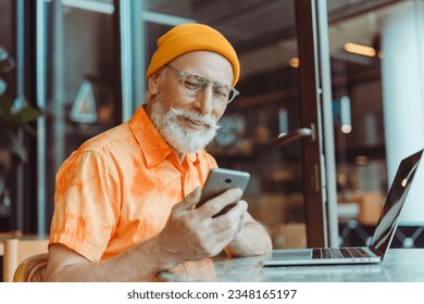 Smiling senior man sitting in modern office, cafe holding mobile phone using laptop checking mail. Elderly hipster wearing stylish yellow hat, shirt and glasses working online. Online shopping concept - Powered by Shutterstock