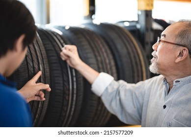 Smiling Senior Man With Spectacles And Beard Checking New Car Tires From Rack For Maintenance And Change In Garage While Getting Assistance From Young Male Mechanic