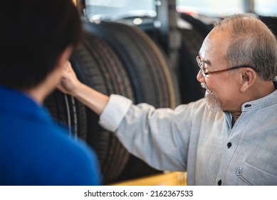 Smiling Senior Man With Spectacles And Beard Checking New Car Tires From Rack For Maintenance And Change In Garage While Getting Assistance From Young Male Mechanic