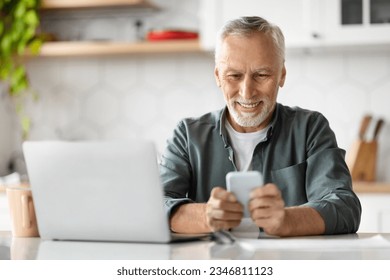 Smiling Senior Man Using Smartphone And Laptop Computer At Home, Happy Elderly Gentleman Texting Via Cellphone And Browsing Internet, Sitting At Desk In Kitchen Interior, Closeup Shot