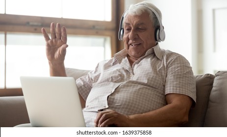Smiling senior man wear earphones wave to camera having video call on laptop, happy elderly male in headphones sit on couch at home talk using modern technologies and wireless connection