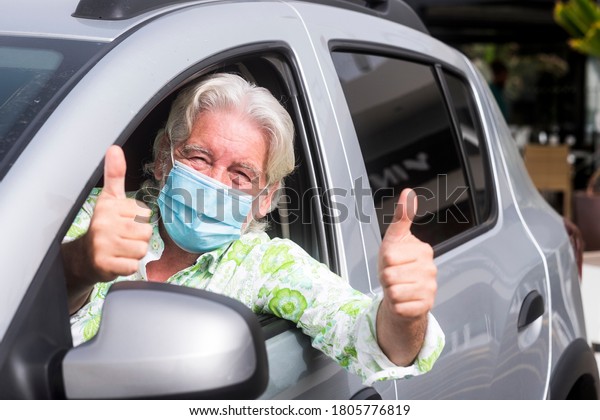 Smiling senior man wearing surgical mask due to\
covid19 coronavirus parked with his silver car gesturing ok sign\
with both hands outside the\
window