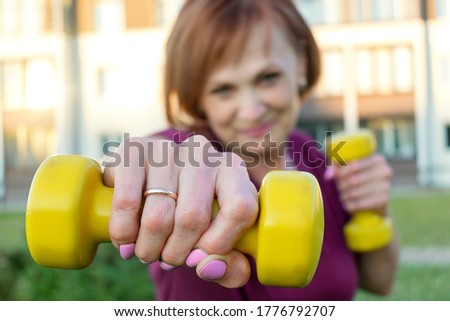 Smiling senior sporty woman exercises with yellow dumbbells in her hands on the front lawn.Selective focus on yellow dumbbells.Woman in a blur.Concept of active lifestyle and healthcare in mature age.