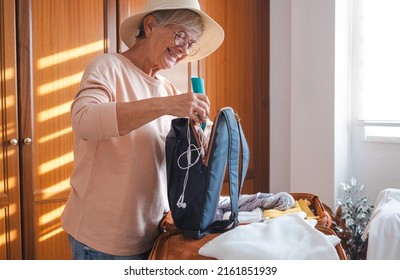 Smiling Senior Tourist Woman Packing Her Accessories Into A Backpack Preparing Luggage For Summer Vacation Trip. Traveling Preparation Concept