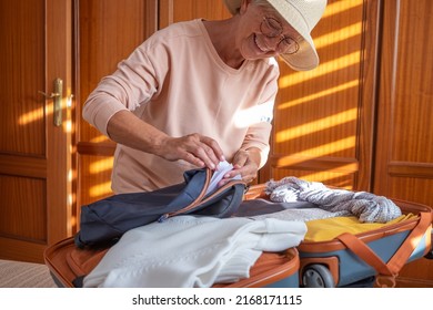 Smiling Senior Tourist Woman Packing Her Accessories Into A Backpack Preparing Luggage For Summer Vacation Trip. Traveling Preparation Concept