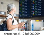 Smiling senior woman in airport looking at timetable schedule to check flight departure gate. Travel and tourism concept, people with backpack on shoulders.