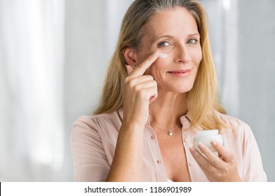 Smiling senior woman applying anti-aging lotion to remove dark circles under eyes. Happy mature woman using cosmetic cream to hide wrinkles below eyes. Lady using day moisturizer.