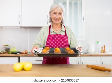 Smiling Senior Woman In Apron Holding Sheet Pan With Just Baked Cupcakes. Old Woman Baking Cakes At Home.