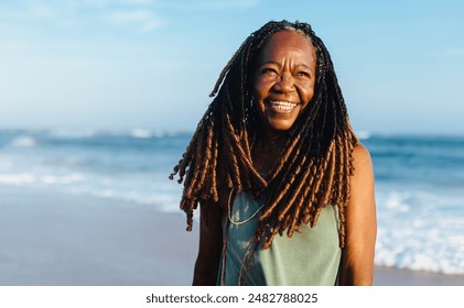 Smiling senior woman with braids standing casually on the beach during sunset. Feeling peaceful and blissful. - Powered by Shutterstock