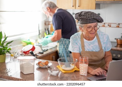 Smiling Senior Woman Browsing By Laptop Looking For A Recipe To Prepare Her Homemade Plumcake While Her Husband Washes The Dishes. Concept Of Family And Domestic Partnership