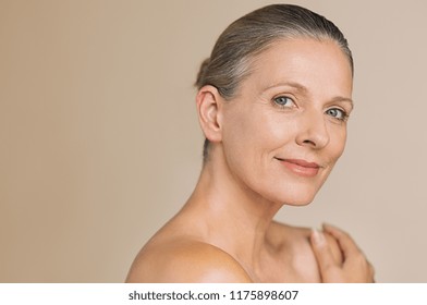 Smiling senior woman with bun hair and hand on naked shoulder. Portrait of beauty mature woman isolated over grey background with copy space looking at camera. Body and skin care concept.