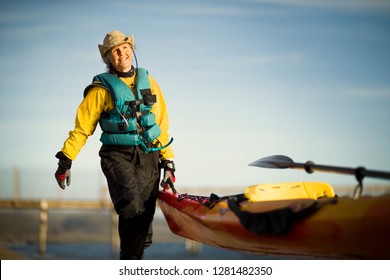 Smiling Senior Woman Carrying The End Of A Kayak In The Sunshine.