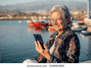 Smiling senior woman in casual jacket using mobile phone while sitting at the sea port at sunset light. Relaxed elderly lady enjoying free time, travel or retirement - Powered by Shutterstock