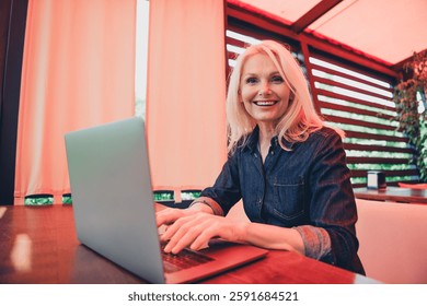 Smiling senior woman in denim jacket using laptop outdoors on a bright day