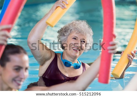 Smiling senior woman doing aqua fitness with swim noodles. Happy mature healthy woman taking fitness classes in aqua aerobics. Healthy old woman holding swim noodles doing aqua gym with young trainer.