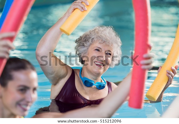Smiling senior woman doing aqua fitness with\
swim noodles. Happy mature healthy woman taking fitness classes in\
aqua aerobics. Healthy old woman holding swim noodles doing aqua\
gym with young trainer.