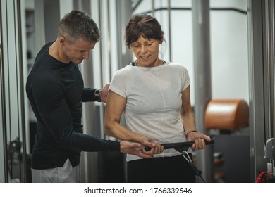 A Smiling Senior Woman Is Doing Exercises On Cable Machine With Personal Trainer In The Gym. They Are Focused On Doing Cable Triceps Pulldowns During A Strength Training Workout.