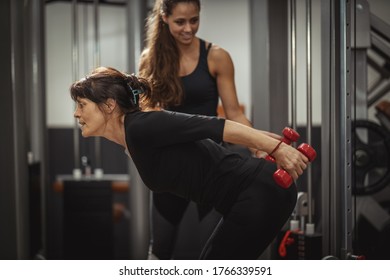 A Smiling Senior Woman Is Doing Exercises With Female Personal Trainer In The Gym. They Are Focused On Doing Triceps Exercises With Dumbbells During A Strength Training Workout .