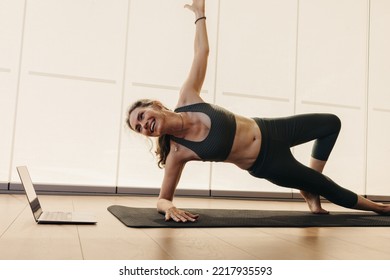 Smiling Senior Woman Doing A Side Plank During An Online Yoga Class. Elderly Woman Practicing A Stretching Asana On An Exercise Mat. Happy Woman Following An Online Workout Video On A Laptop.