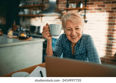 Smiling senior woman drinking coffee and using laptop at home - Powered by Shutterstock