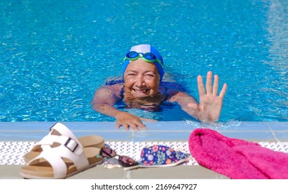 Smiling senior woman at the edge of the swimming pool greetings with hand, wearing swimming cap and goggles. Happy elderly lady enjoying healthy lifestyle and holidays