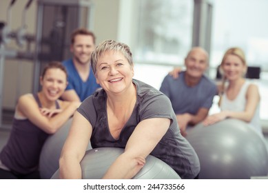 Smiling Senior Woman Enjoying Pilates Class At The Gym Posing Leaning On Her Ball Smiling At The Camera With The Class Behind