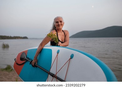 a smiling senior woman with gray hair holding sup board on the seashore at sunset. copy space. Slow life. Enjoying the little things. spends time in nature in summer - Powered by Shutterstock