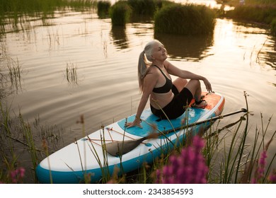 a smiling senior woman with gray hair sitting on sup board on the seashore at sunset. copy space. Slow life. Enjoying the little things. spends time in nature in summer - Powered by Shutterstock