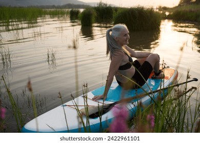 a smiling senior woman with gray hair sitting on sup board on the seashore at sunset. copy space. Slow life. Enjoying the little things. spends time in nature in summer - Powered by Shutterstock