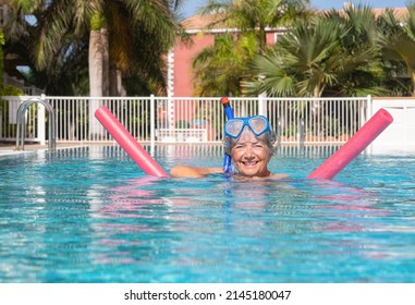 Smiling senior woman have fun in swimming pool with swim noodles and diving mask. Happy mature healthy woman taking fitness classes in aqua aerobics.