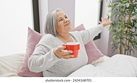 A smiling senior woman holding a red mug, stretching in bed with pink pillows and a plant in a cozy bedroom - Powered by Shutterstock