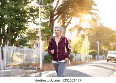 Smiling Senior Woman Jogging On Streets In Early Morning. Active Mature Woman Running On A Sunny Day While Listening To Music. Lady Running With Armband And Listening To Music At Sunset.