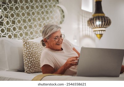 Smiling senior woman lying in bedroom at home or hotel room with laptop using mobile phone enjoying tech and social. - Powered by Shutterstock