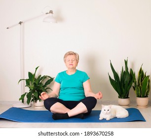 Smiling Senior Woman Meditating Relaxing With Her White Cat On Fitness Mat. Granny In Yoga Pose Doing Breathing Exercises At Home. Old Age, Tranquility, Health And Life Style Concept.