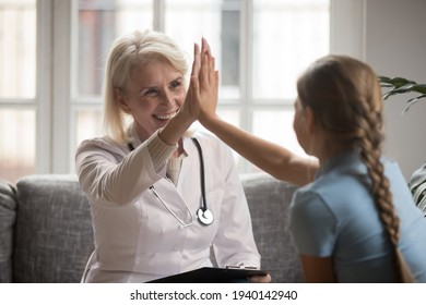 Smiling Senior Woman Nurse Or GP Give High Five Celebrate Good Results With Teen Girl Kid In Clinic. Happy Mature Female Doctor Or Pediatrician Make Deal With Small Child Patient. Healthcare Concept.