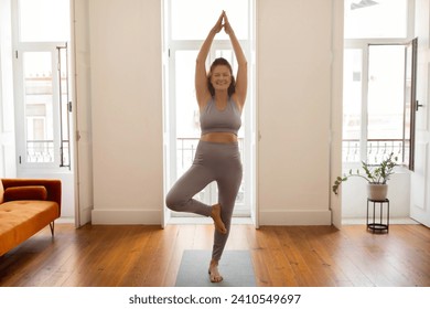 Smiling senior woman practicing Tree pose with hands in Anjali Mudra, active elderly lady training yoga at home, exuding balance and peace, exercising in sunlit living room interior, copy space - Powered by Shutterstock