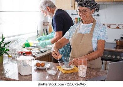 Smiling Senior Woman Preparing Her Homemade Plumcake While Her Husband Washes The Dishes. Concept Of Family And Domestic Partnership